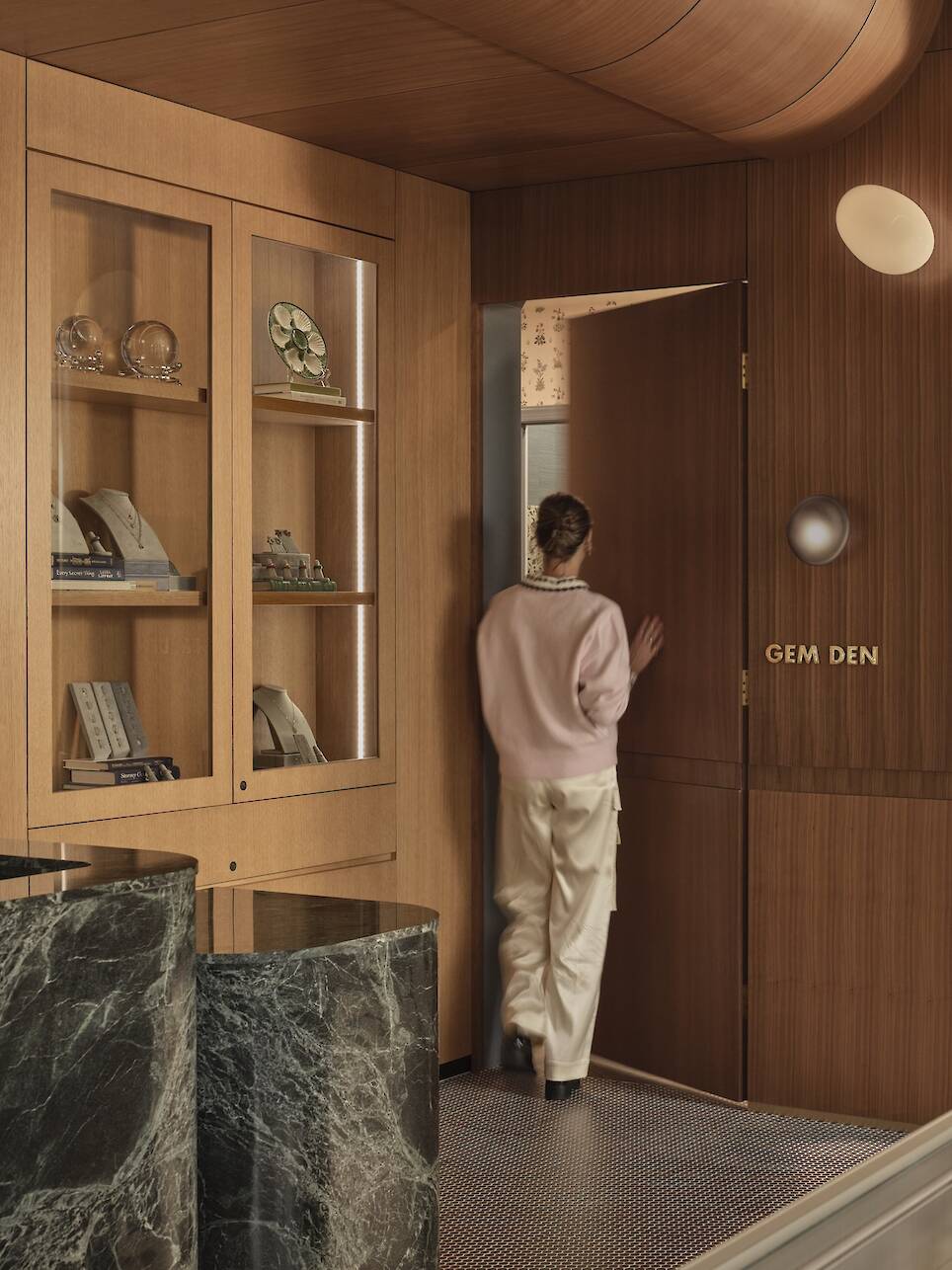 A person in a pink sweater and white pants enters a wooden door labeled "GEM DEN" in a room with wood paneling, marble counters, and display shelves.
