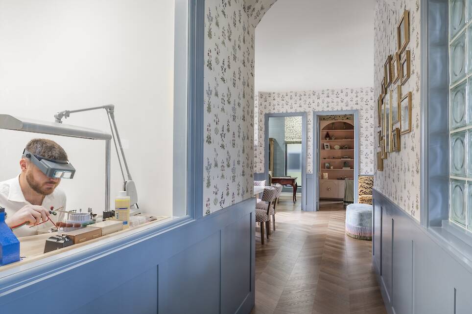 A person wearing magnifying goggles works on a small project at a desk in a room with floral wallpaper and blue wainscoting, viewed from a hallway leading to a furnished living...