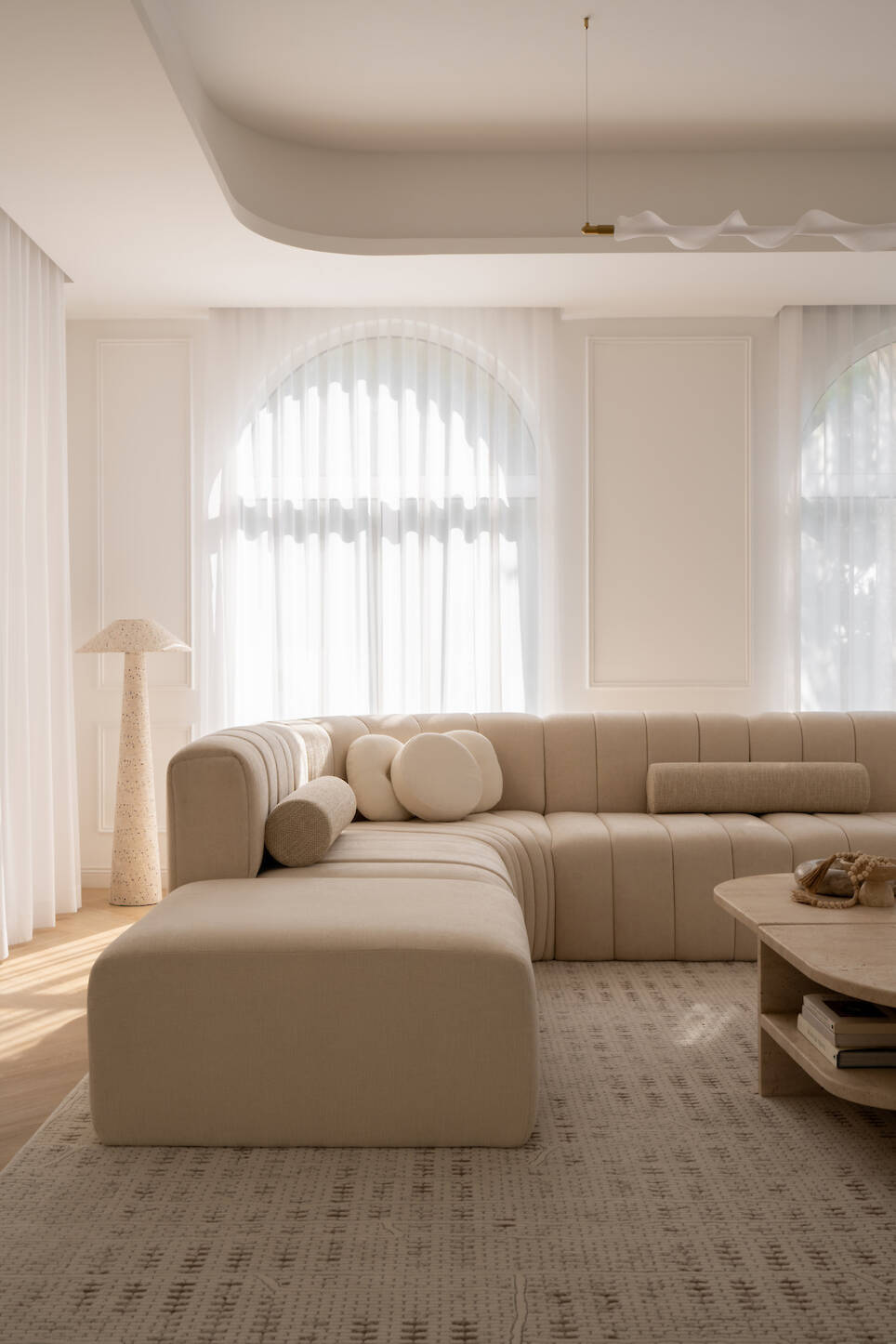 Modern living room with a beige sectional sofa, round cushions, textured rug, wooden coffee table, floor lamp, and large arched windows with sheer curtains