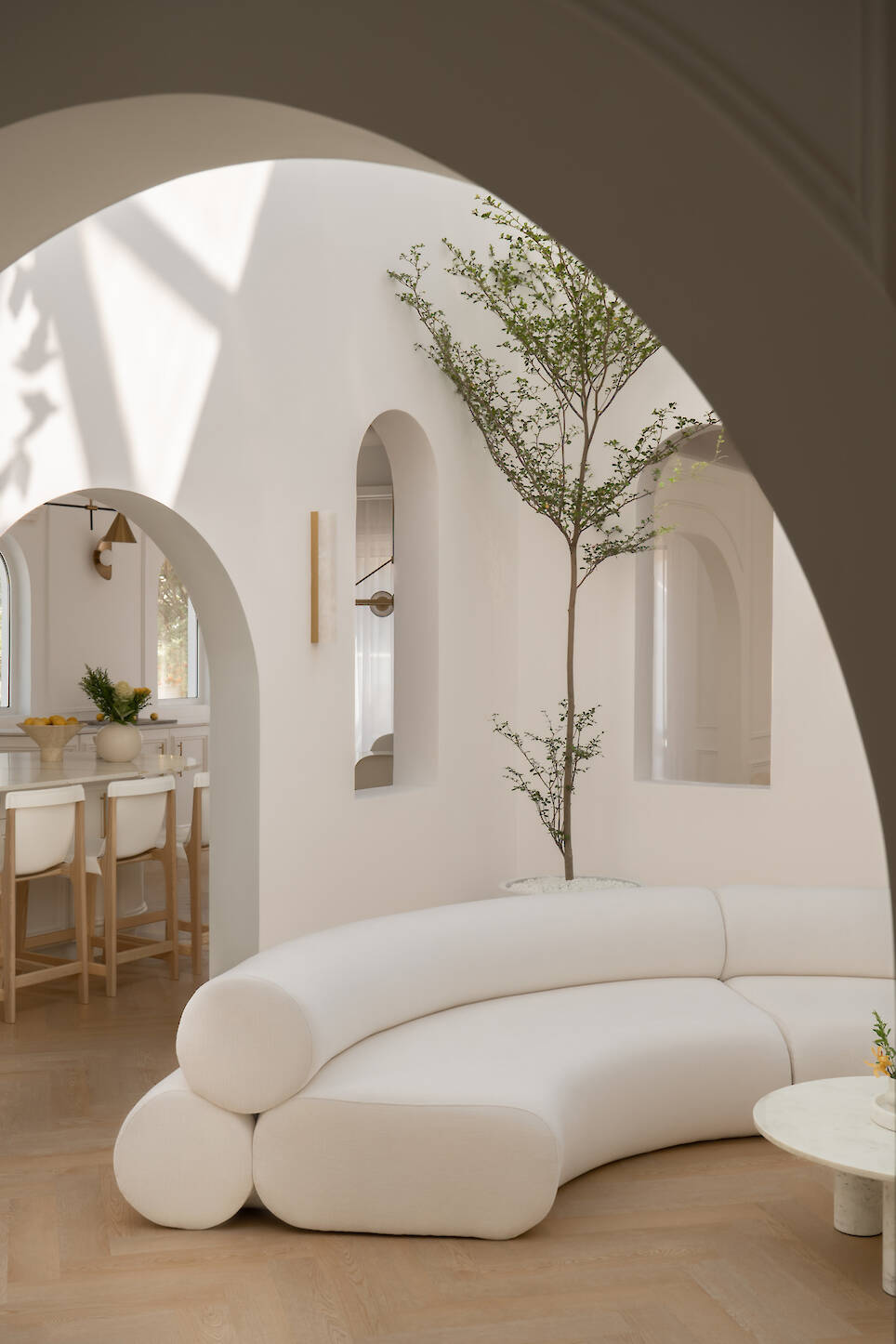 Minimalist living room with curved white sofa, a round coffee table, arched doorways, indoor tree, and light wood floor. Sunlight casts shadows on white walls