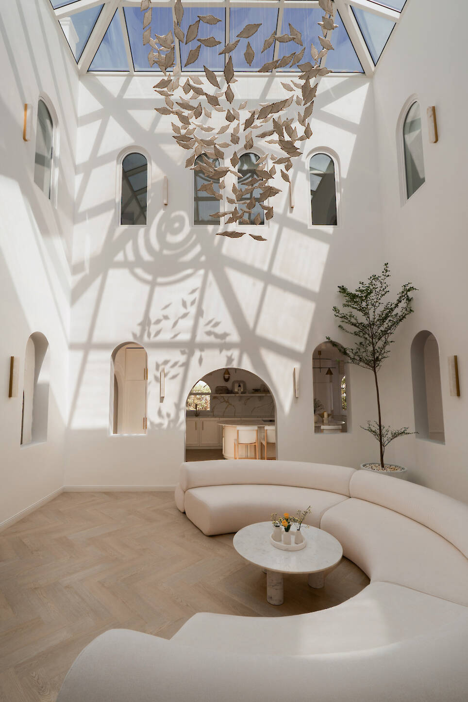 A bright, modern atrium by D’Ora Tokai Designs features white curved seating, a round table, skylight, abstract hanging sculpture, large windows, and a potted tree on a light
