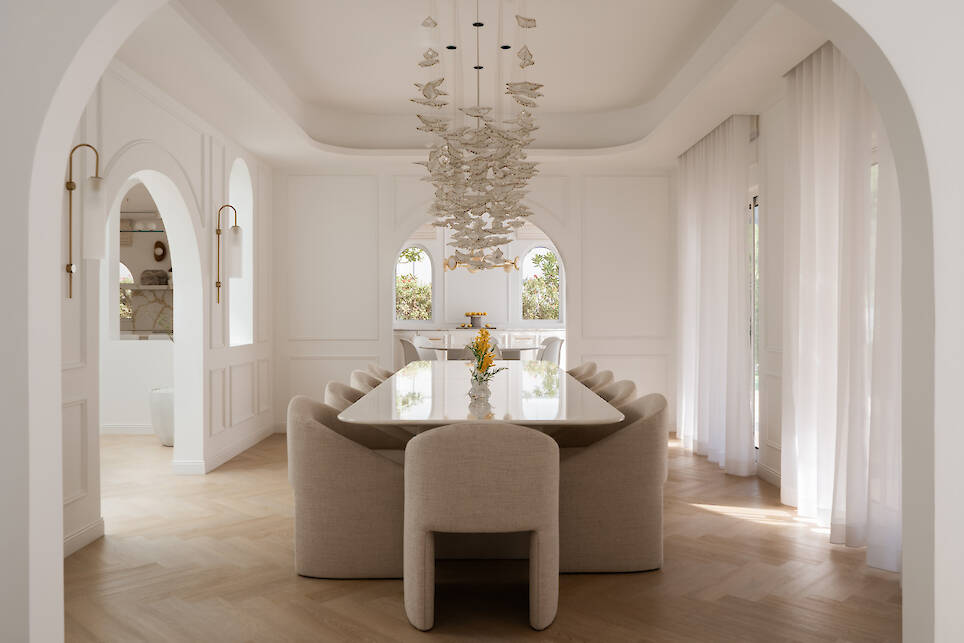 Modern dining room with a long table, beige upholstered chairs, sculptural chandelier, arched doorways, light wood floors, and sheer white curtains letting in natural light