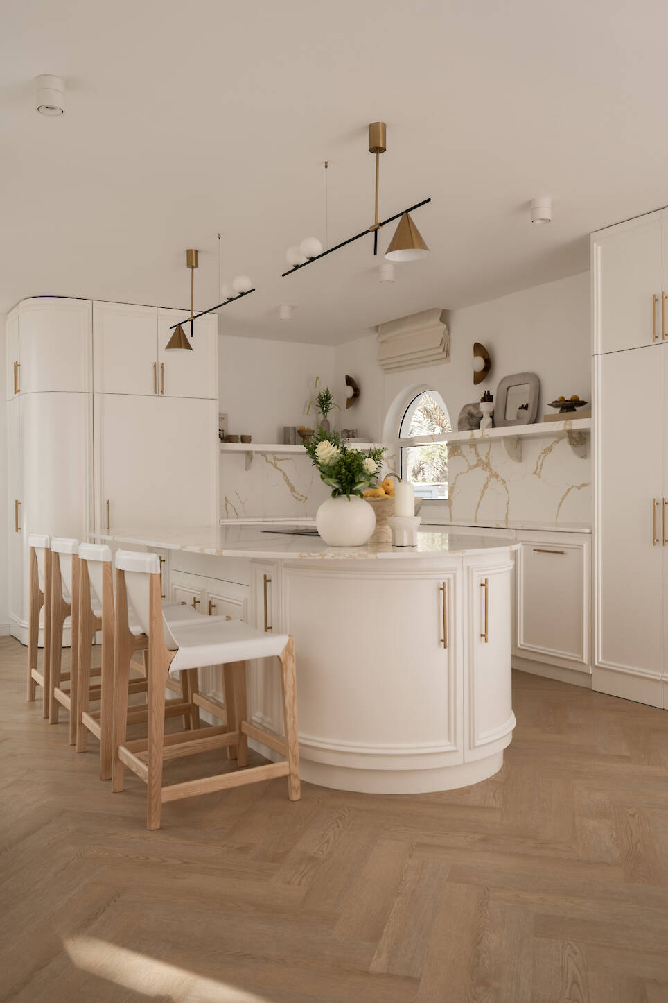 Modern kitchen with white cabinets, marble backsplash, a curved island with four wooden stools, gold fixtures, and neutral décor. Natural light enters through an arched window