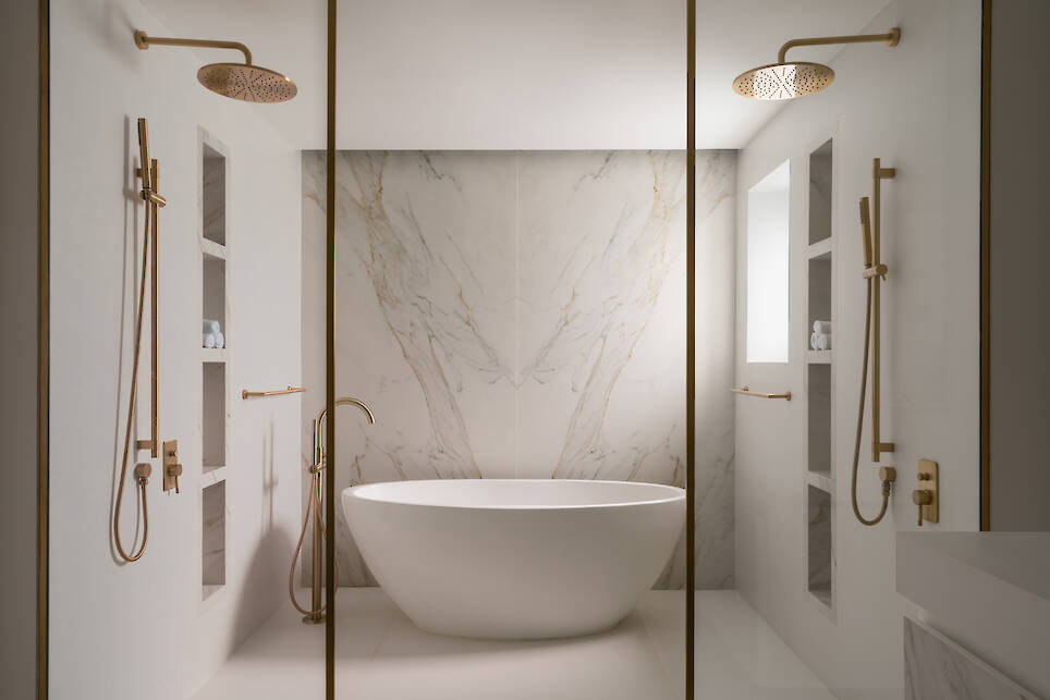 Modern bathroom with a freestanding oval bathtub, marble accent wall, dual gold rain showers, built-in shelves, and white walls