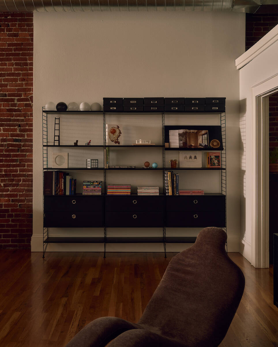 Black metal bookshelf with drawers, books, decorative items, and boxes stands against a white wall with exposed brick and wooden floor. A brown chair is partially visible in the