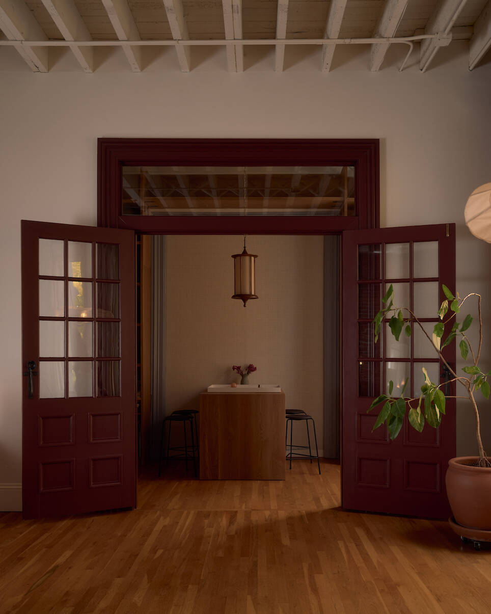 Open double doors reveal a small table with two stools in a softly lit room; a potted plant stands to the right on a wooden floor under an exposed beam ceiling