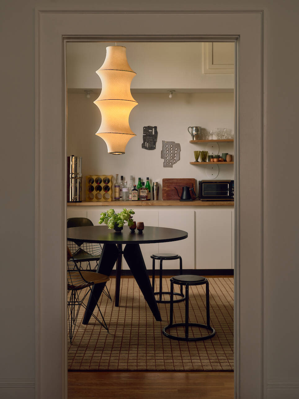 A modern dining area with a round black table, four chairs, a sculptural hanging lamp, and a kitchen with shelves and various items in the background
