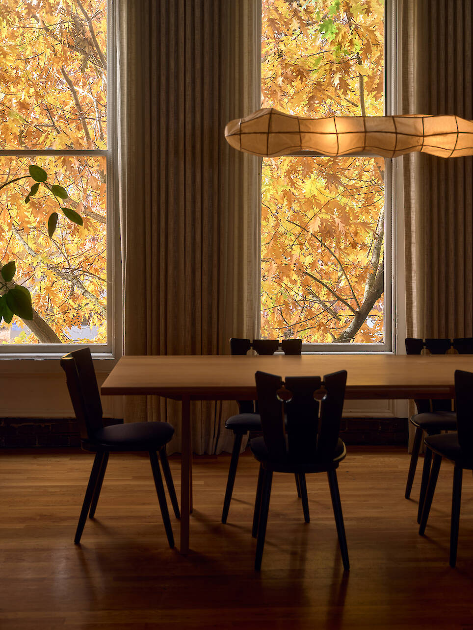 A wooden dining table with black chairs sits by large windows with autumn leaves visible outside; a modern light fixture hangs above