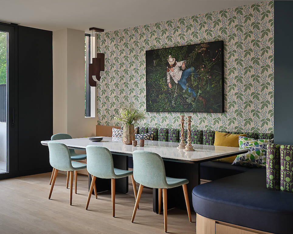A modern dining area with a marble table, light blue chairs, patterned wallpaper, a large plant-themed photo, and built-in bench seating with assorted cushions creates an inviti