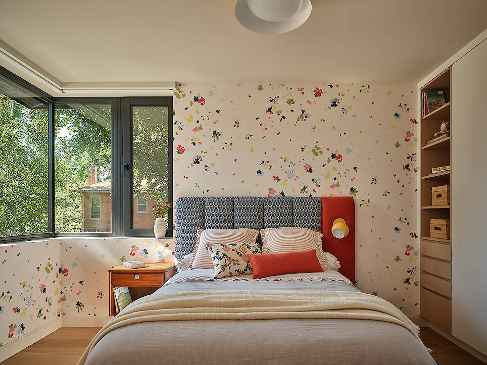 A cozy bedroom with floral wallpaper, a bed with colorful pillows, a wooden nightstand, built-in shelves, and large windows overlooking green trees embodies the thoughtful desig