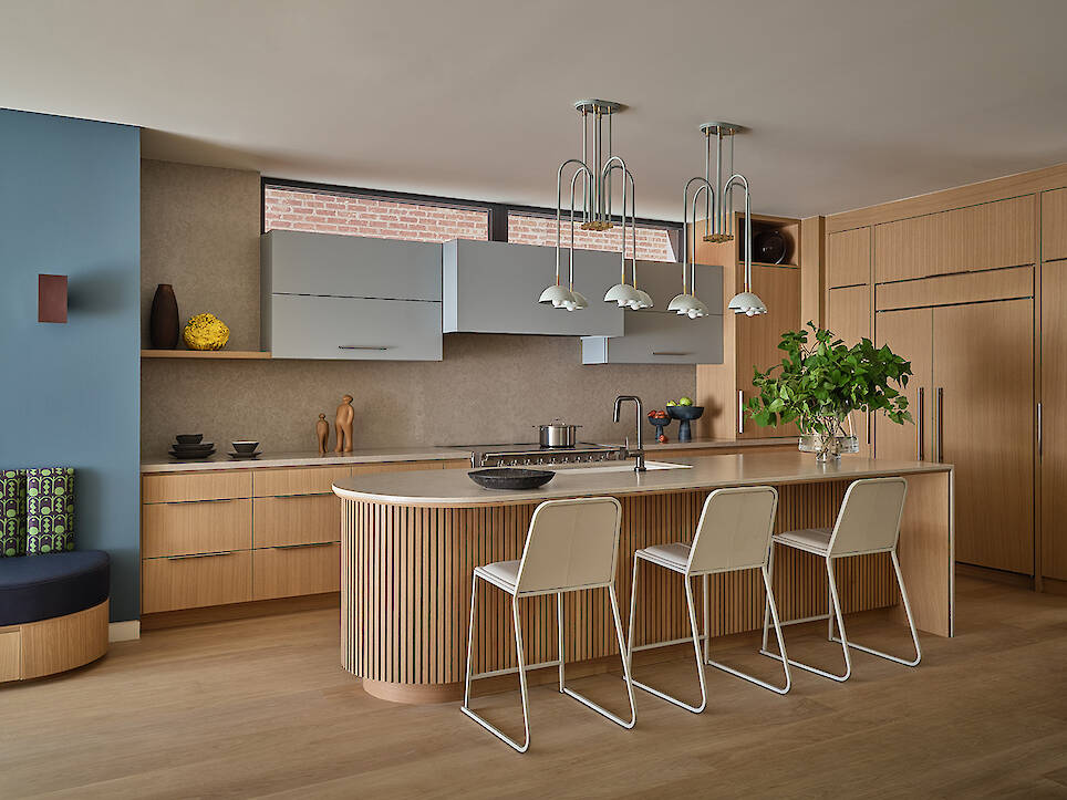 Modern kitchen designed by Gillian Gillies in a Net Zero Ready Home, featuring light wood cabinets, a curved island with three white barstools, blue accent wall, and pendant lig
