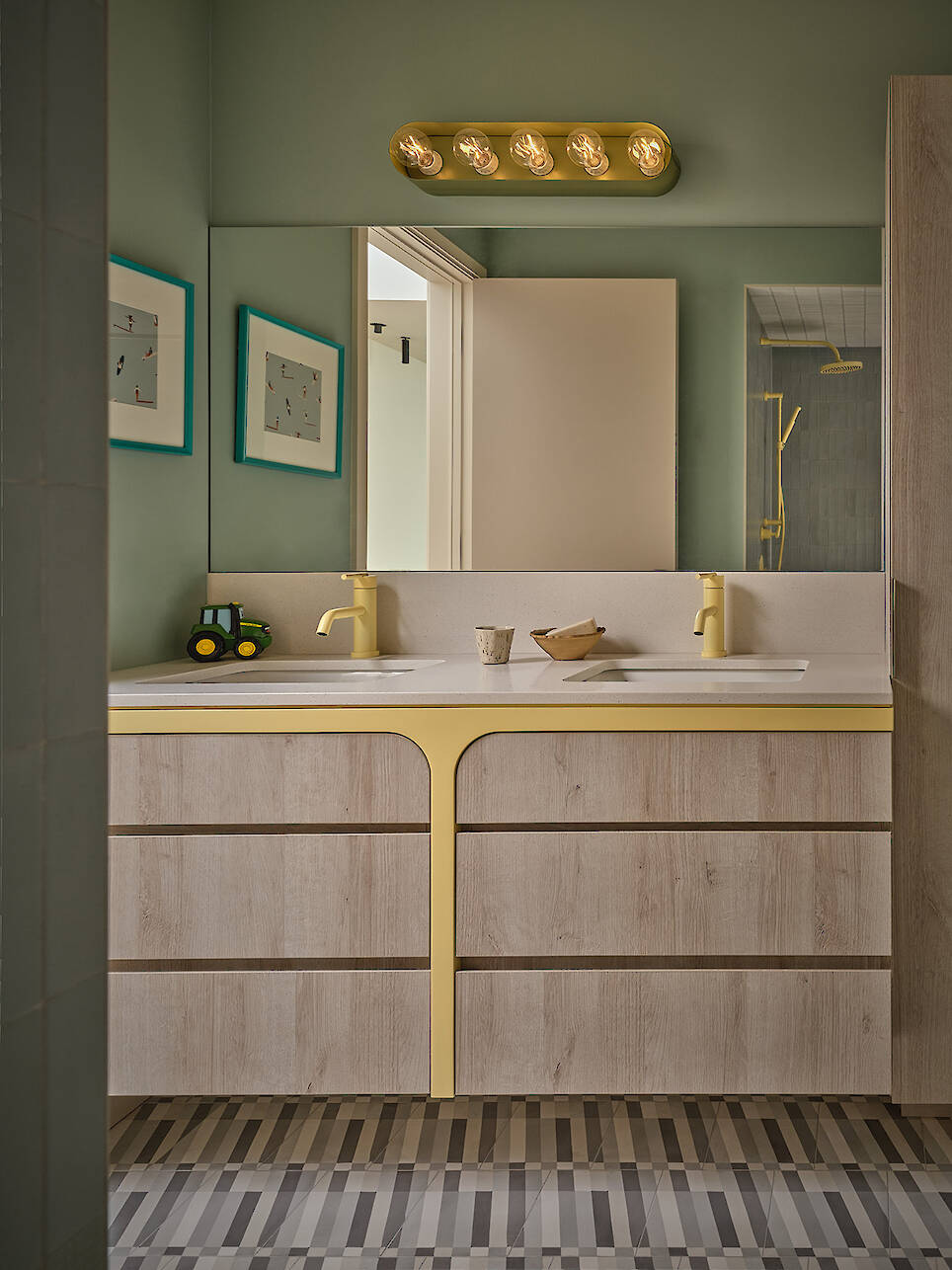 Bathroom in a Gillian Gillies Net Zero Ready Home with dual sinks, light wood vanity, yellow accents, large mirror, wall-mounted lights, green walls, framed art, and a toy tract