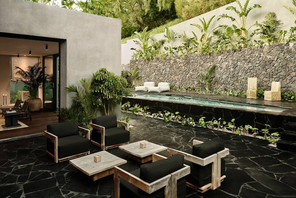 Modern outdoor patio with black stone floor in an elegant Obsidian House style, wooden furniture with black cushions, a small pool, lush plants, and a beautiful stone wall