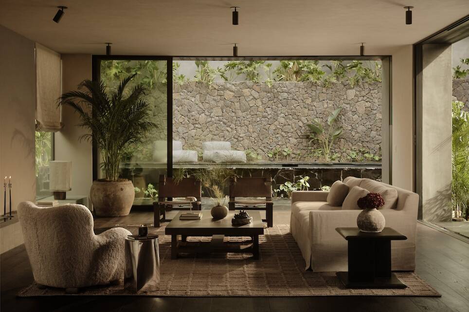 Modern living room with minimalist furniture, large windows, and a view of a stone wall and lush greenery outside evoke the serene elegance of an Obsidian House. Neutral tones a
