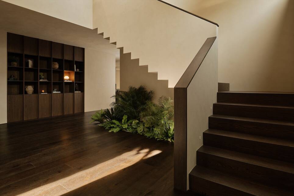 Modern interior of Obsidian House with a dark wooden staircase, built-in shelving, and green indoor plants nestled beneath the stairs. Warm natural light illuminates the dark wo