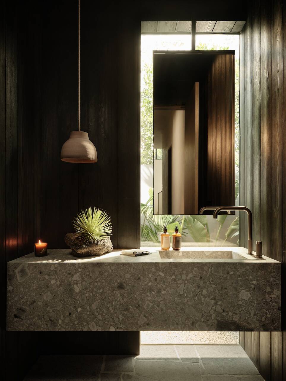 Modern bathroom inside Obsidian House with a stone sink, wall-mounted faucet, decorative plant, candle, toiletries, and a mirror reflecting outdoor greenery. Natural light enter