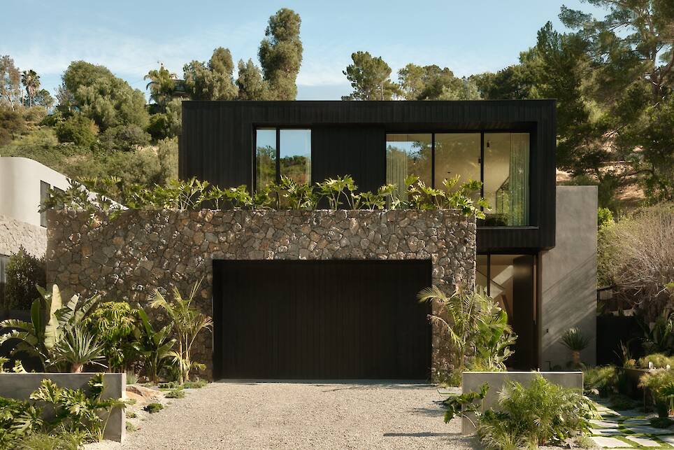 Modern two-story Obsidian House with large windows, stone facade on the lower level, black upper level, surrounded by lush green plants and trees. Gravel driveway leads to a bla