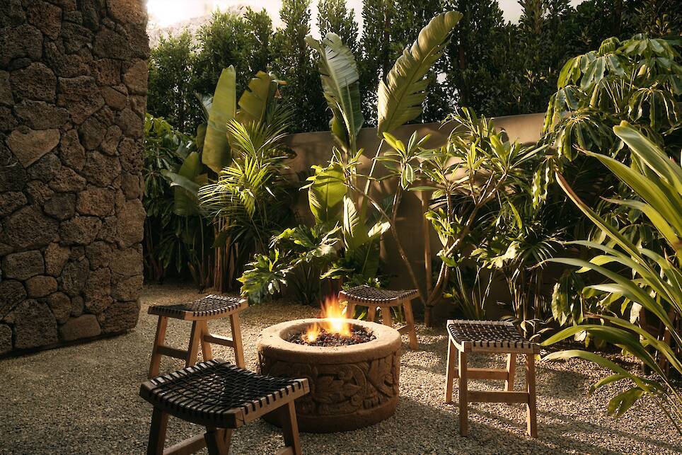 Four woven stools surround a stone fire pit with a small fire, set in the gravel courtyard of Obsidian House, framed by dense tropical plants and a striking stone wall