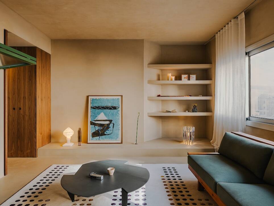 A minimalist living room with beige walls, a green sofa, a dark geometric coffee table on a patterned rug, built-in shelves holding decor, and a framed abstract painting leaning