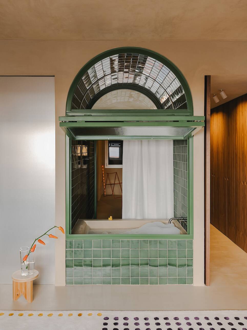 A modern bathtub is set within green tiles beneath an arched window, with a side table holding a glass and orange flowers nearby