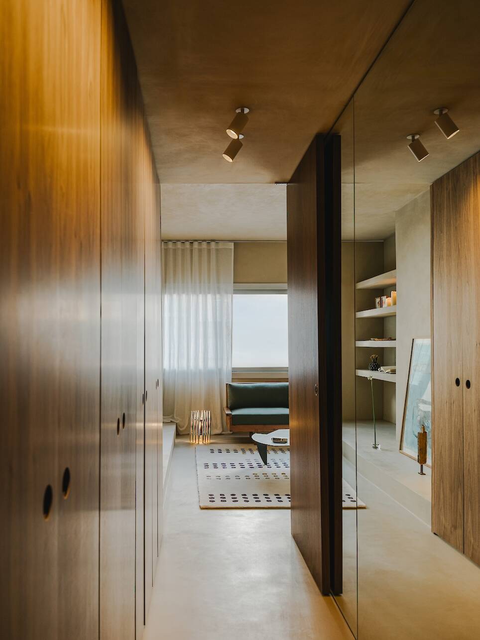 Modern hallway with wooden closets, mirrored wall, and a view into a sitting area with a window, couch, shelves, and a patterned rug