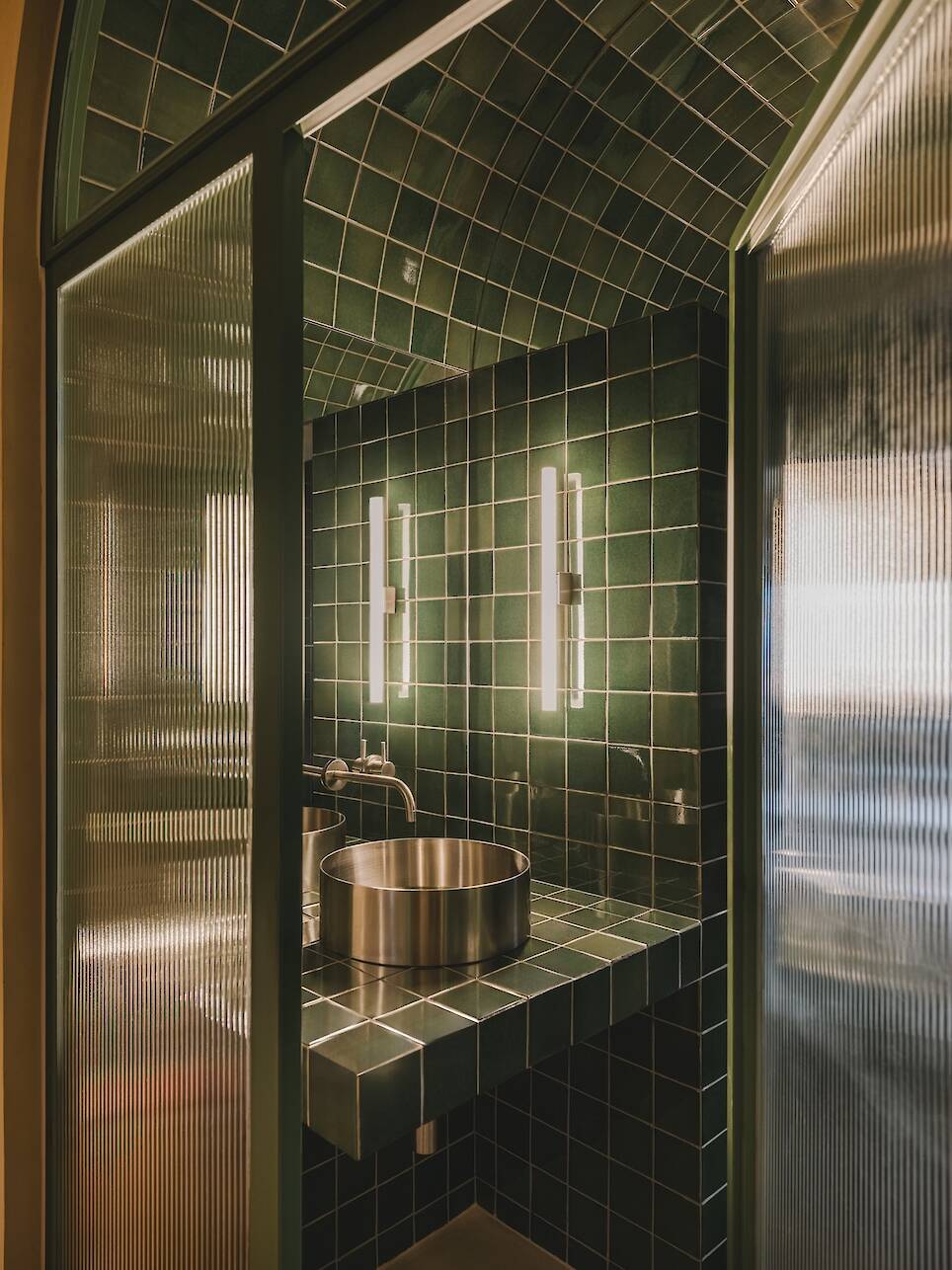 A modern bathroom with green tiled walls and arched ceiling, a stainless steel sink, and vertical wall lights on either side of a mirror