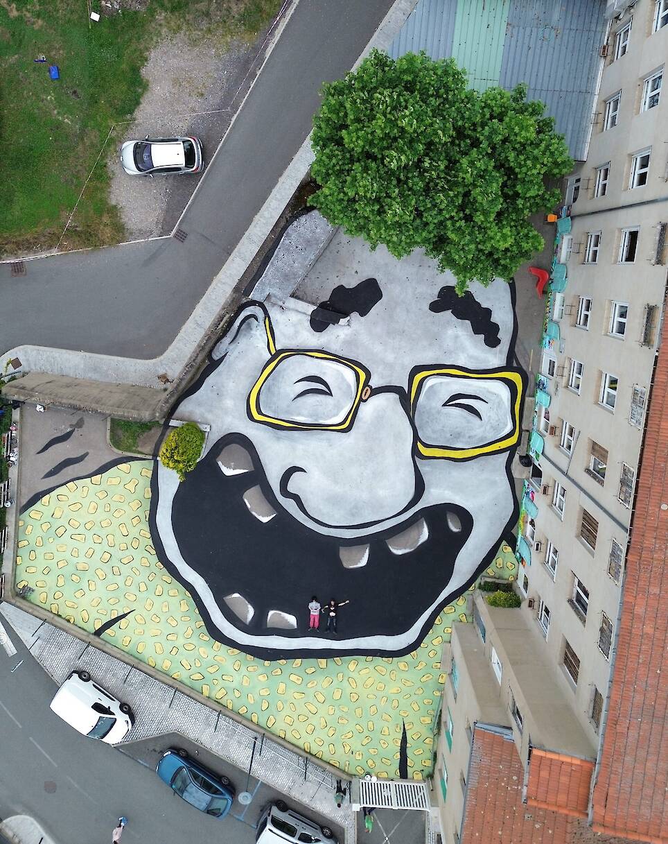 an aerial view of a large mural by Ella & Pitr on the ground of a small courtyard in a neighborhood featuring a cartoonish laughing figure with a tree that looks like hair