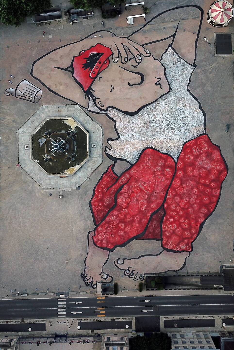 an aerial view of a large mural by Ella & Pitr on the ground of a public square featuring a cartoonish sleeping figure