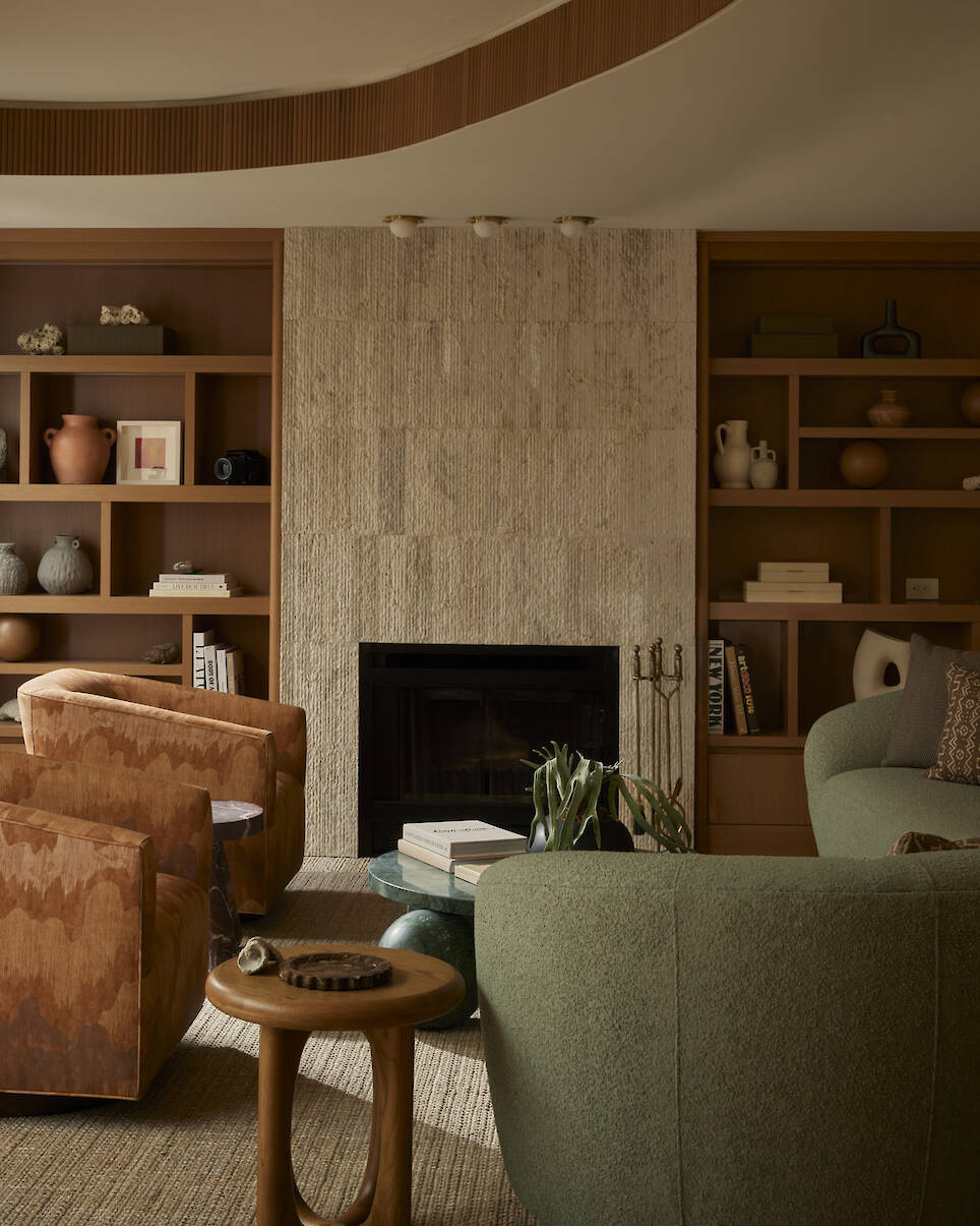 A living room with a stone fireplace, built-in shelves, tan armchairs, a green sofa, and a round side table. Neutral tones and decorative vases create a warm, modern atmosphere