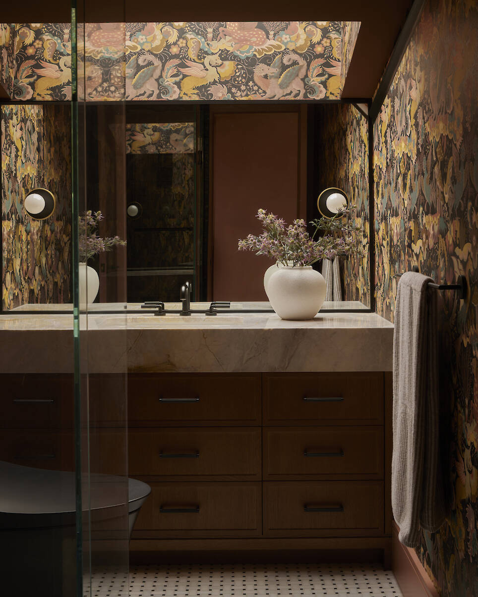 A bathroom with floral wallpaper, a marble countertop, a large white vase with flowers, wall-mounted lights, and a towel hanging on a rack beside the sink