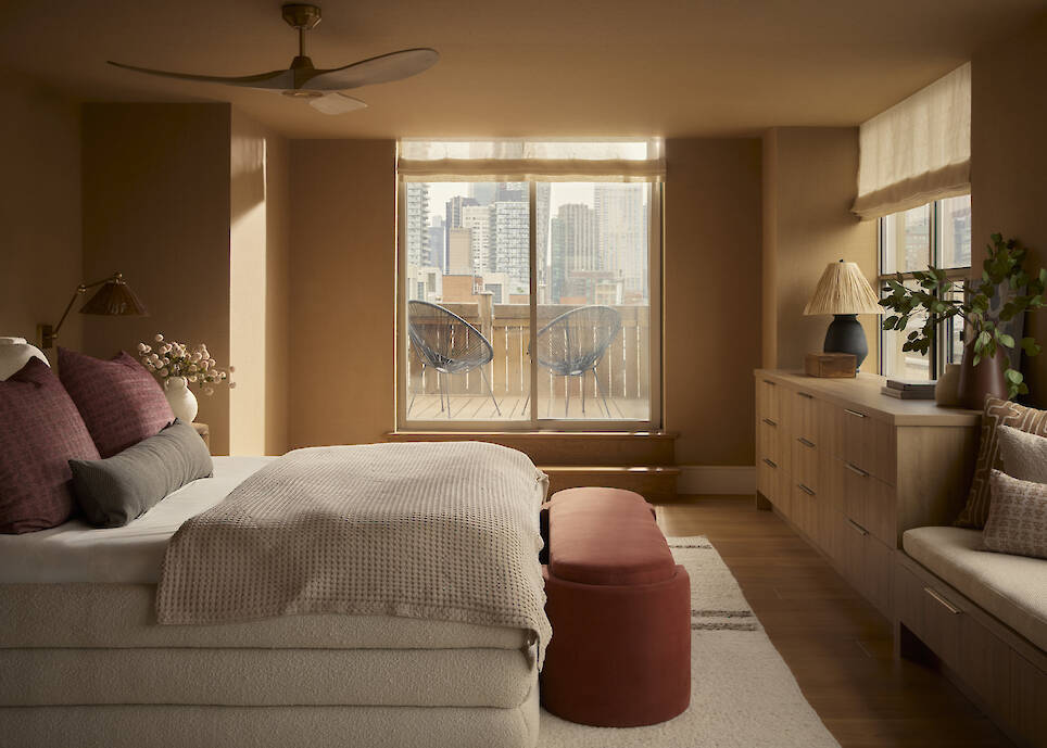 Bedroom with beige walls, bed with pillows, red bench, wooden dresser, large window, city buildings view, and two chairs on the balcony