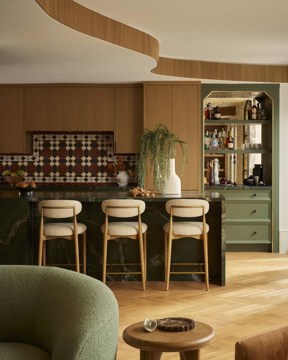 Modern kitchen with wood cabinets, patterned tile backsplash, three bar stools, green accents, a curved ceiling detail, and a bar area with bottles and glassware in the background