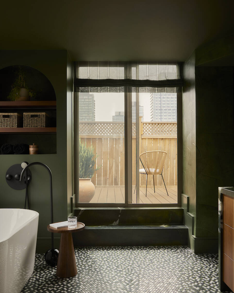 Modern bathroom with a freestanding tub, black fixtures, a small wooden table, dark green walls, and a large window overlooking a balcony with a chair and potted plant