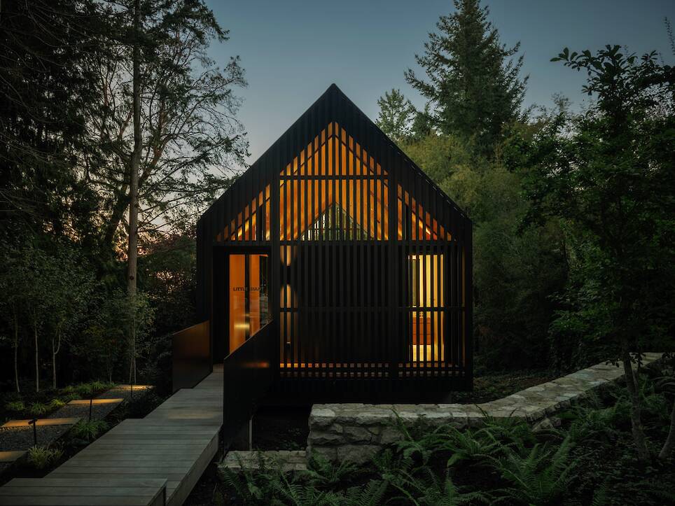 Modern black wood cabin with slatted exterior and warm interior lighting, the Little Giant forest studio is surrounded by trees and greenery at dusk, viewed from a raised walkway