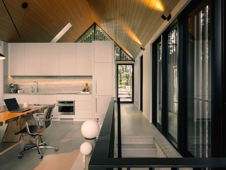 Modern kitchen with white cabinets, built-in appliances, a wooden dining table, and an office chair beside large windows and a sloped wooden ceiling—reminiscent of the cozy cr