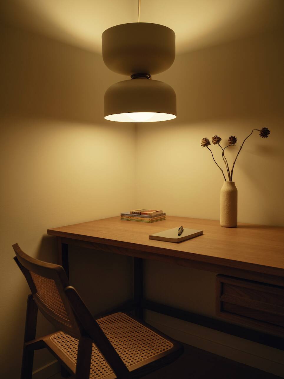 A wooden desk with a notebook, pen, books, and a vase with dried flowers rests under a modern pendant lamp, creating the warm ambiance of a Little Giant forest studio. A woven c