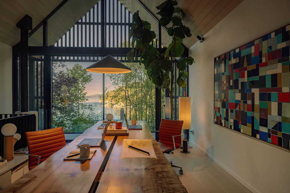 Modern home office with two desks, orange chairs, large windows showing trees and a lake at sunset, a tall plant, and a colorful abstract painting on the wall—this inviting sp