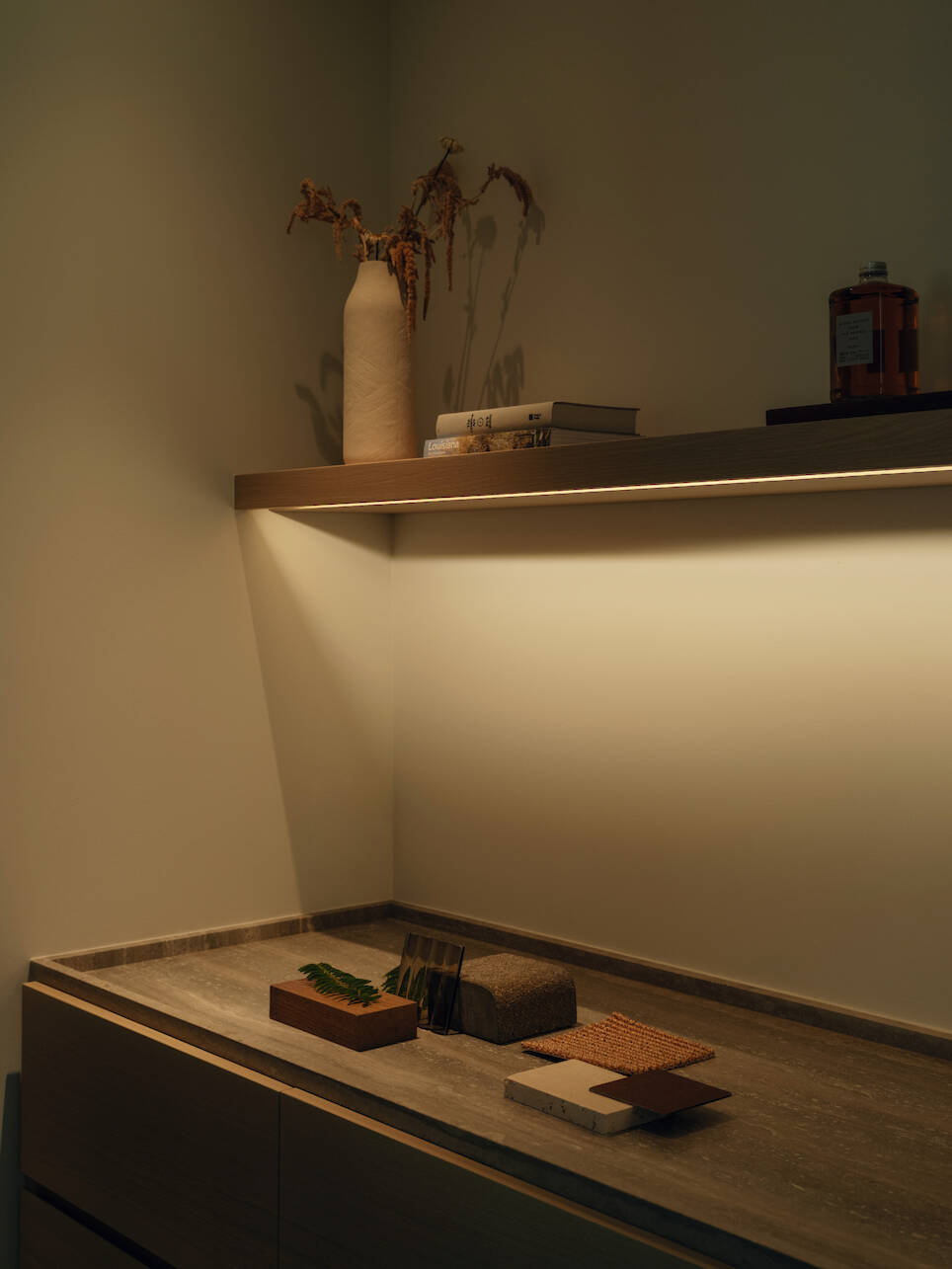A minimalist countertop by Little Giant forest studio features samples and greenery, sitting beneath a lit floating shelf that holds a vase with dried plants, books, and a brown