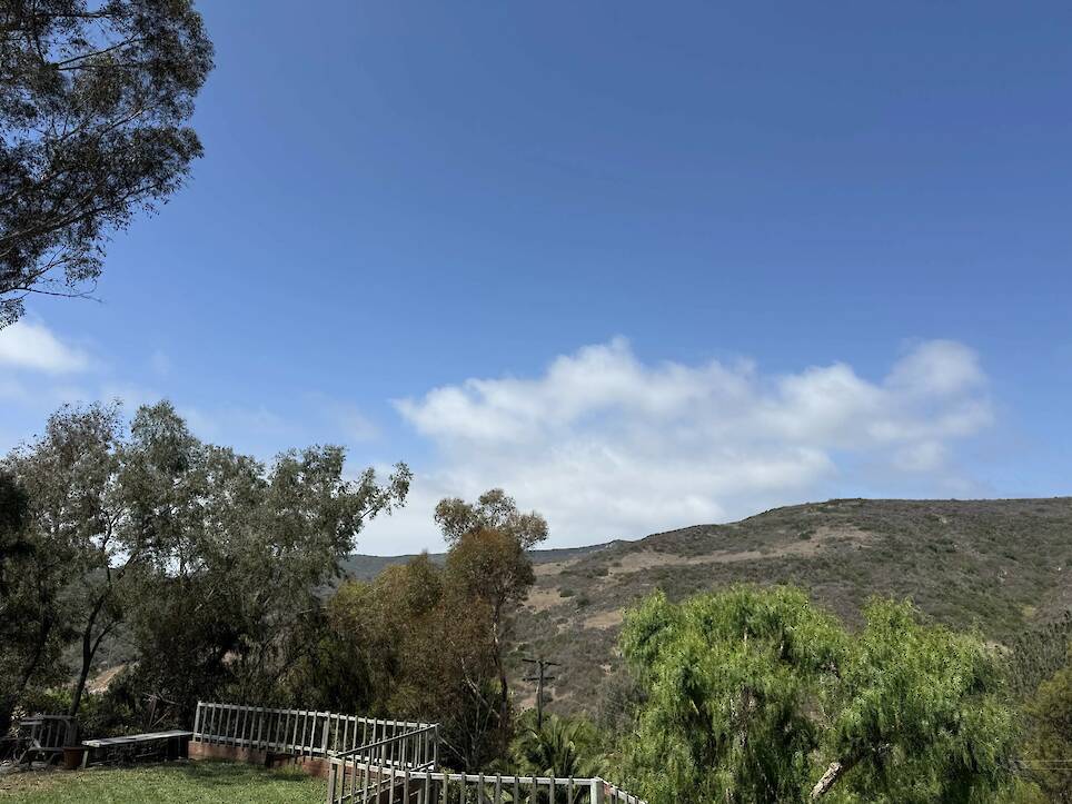 A clear sky with scattered clouds above green hills and trees, with a white fence in the foreground