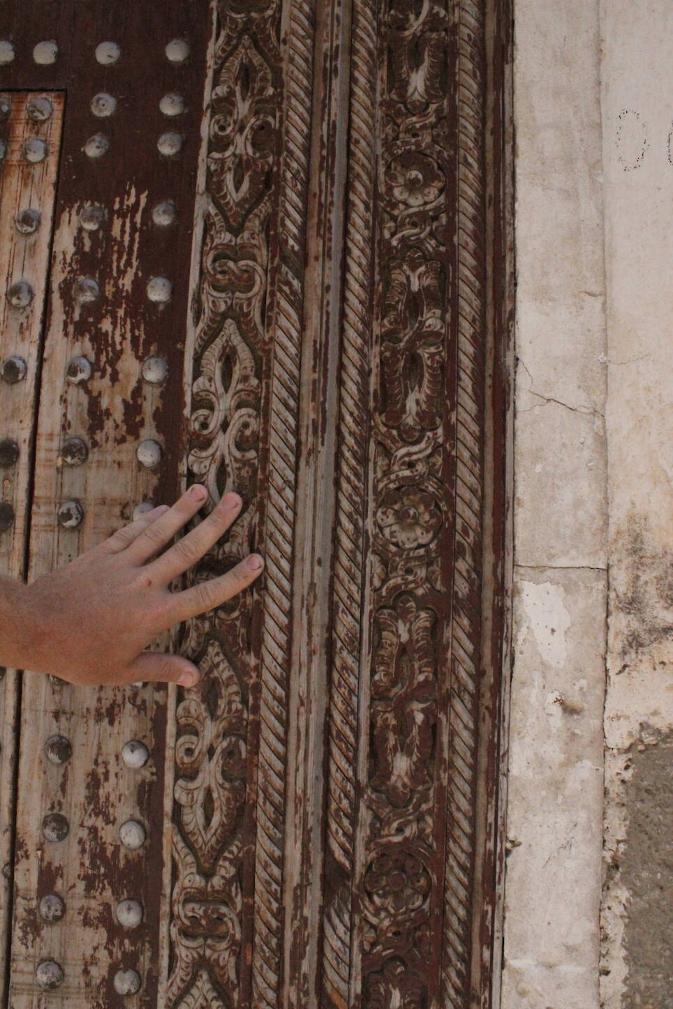 A hand touches an ornate, weathered wooden door with intricate carvings, metal studs, and a stone wall on the right side