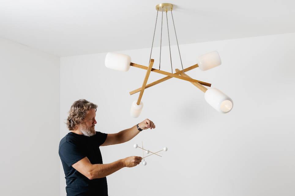 A man holding a small model of a modern chandelier—reminiscent of a Nick Sheridan Cerno design—stands under a large, similarly styled chandelier in a pristine white room
