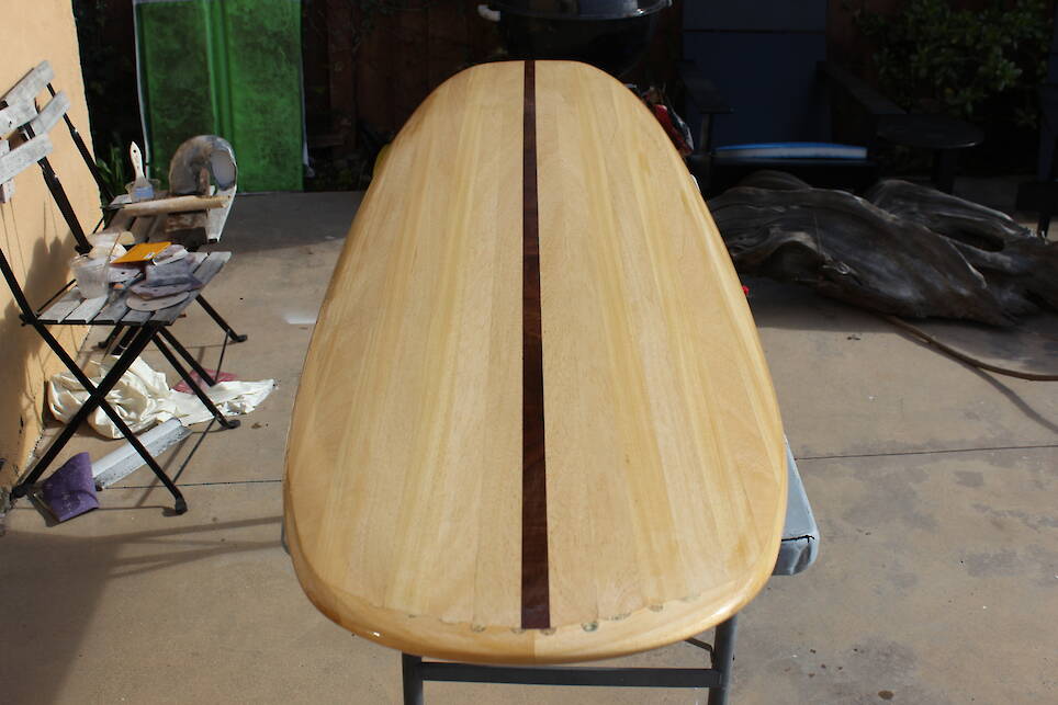 A wooden surfboard with a dark center stripe rests on sawhorses in an outdoor workspace, surrounded by tools and chairs