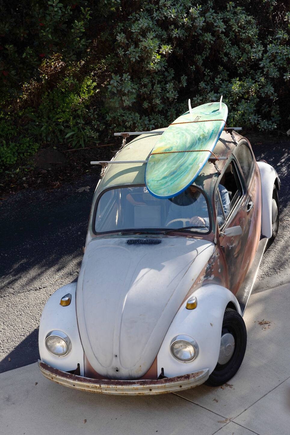Vintage Volkswagen Beetle with worn paint parked on a driveway, carrying two surfboards on its roof
