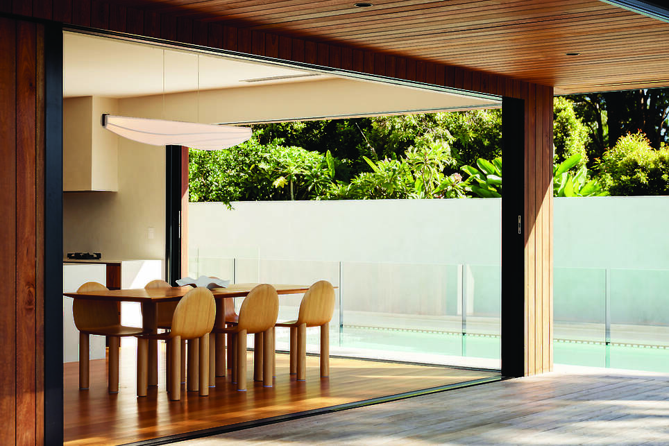 A modern dining area with a wooden table and chairs, illuminated by a sleek Nick Sheridan Cerno light fixture, opens to a poolside patio surrounded by greenery and a white priva