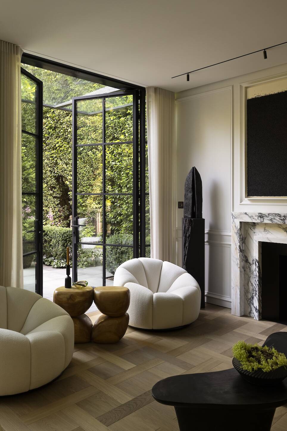 Modern living room with curved white chairs, marble fireplace, abstract art, sculptural tables, and large glass doors opening to a green garden