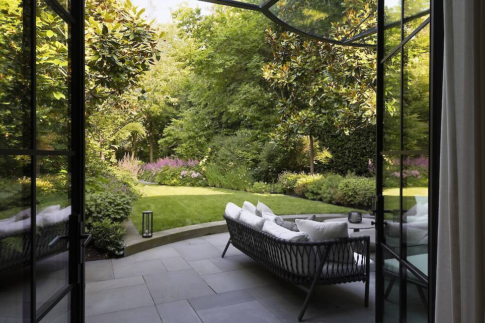 Glass doors open to a patio with outdoor seating, overlooking a landscaped garden with lush greenery, trees, and flowering plants