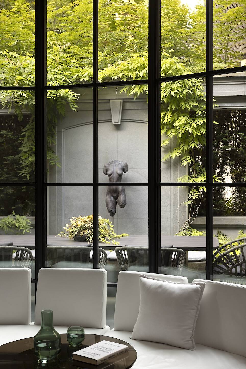 Large windows reveal a garden patio with a classical torso sculpture, lush greenery, patio chairs, and potted plants, viewed from inside a modern living room with white seating