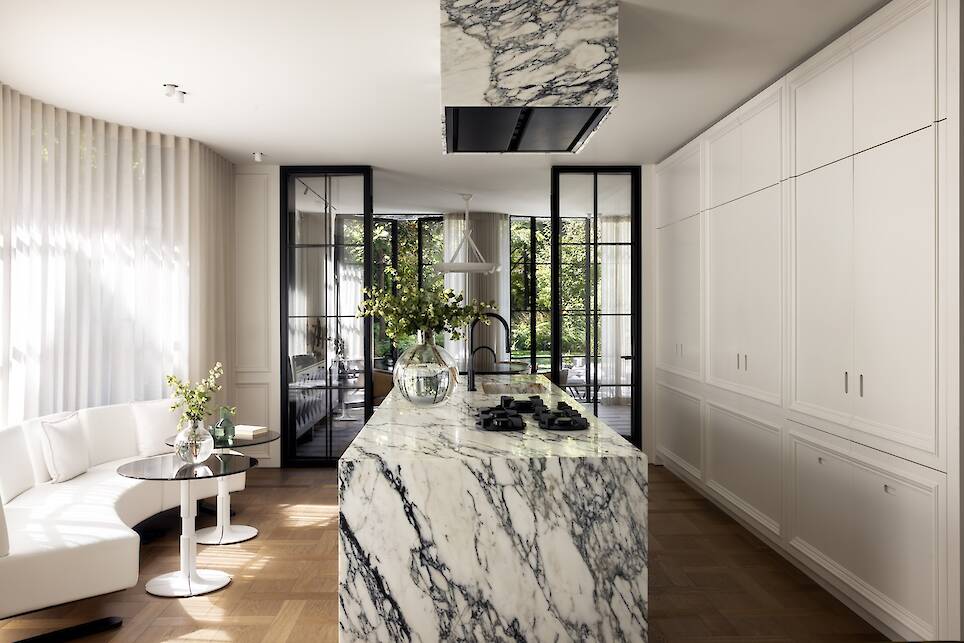 Modern kitchen with a large marble island, white cabinets, glass doors, wood flooring, and a small seating area with a round table and white bench. Sunlight enters through large