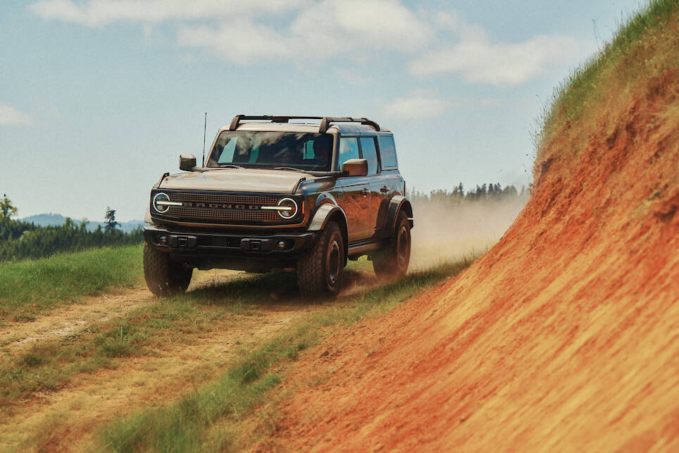 A Ford Bronco drives on a dirt trail next to a steep, orange-colored hillside under a partly cloudy sky