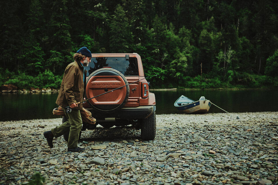 A person carrying a fishing rod walks past an SUV parked on a rocky riverbank, with a boat tied to the shore and a forest in the background